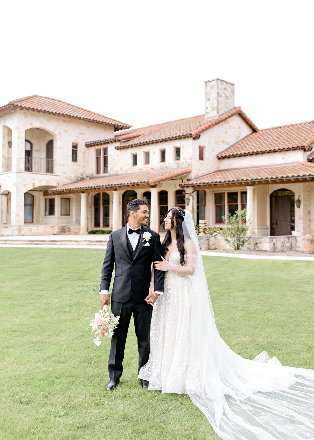 An elegant outdoor wedding ceremony with Spanish-style architecture and a flowing white dress on a grassy lawn.
