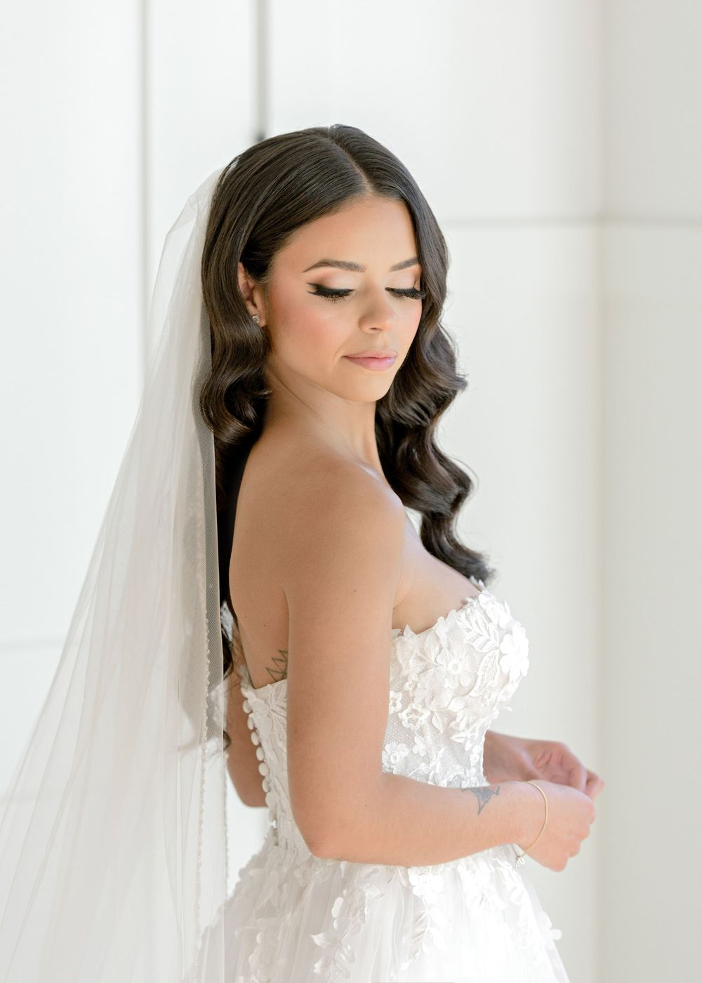 Elegant bride in white lace wedding dress with flowing veil and dark wavy hair styled to one side.
