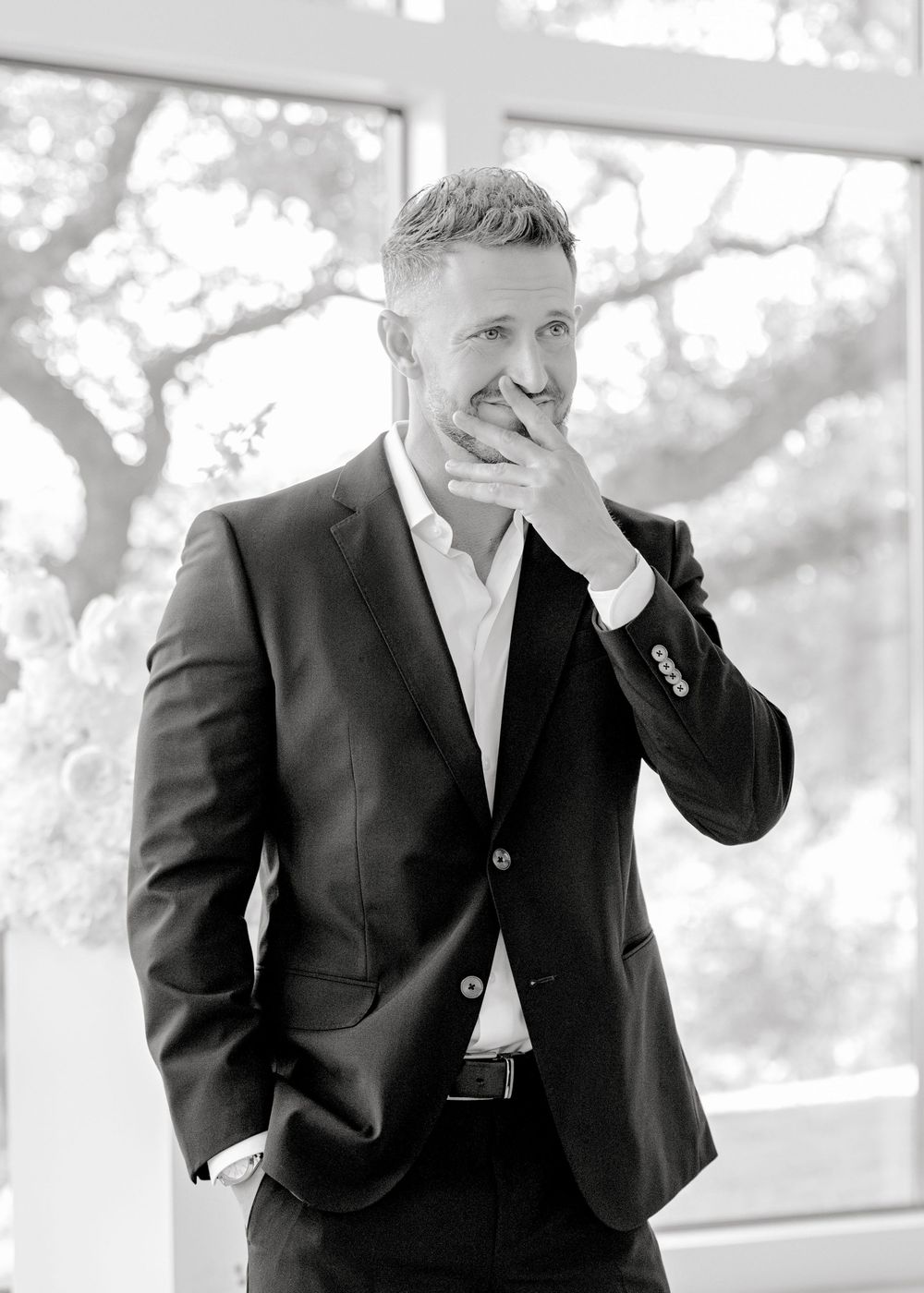 Professional wearing a dark suit poses thoughtfully with hand near face in outdoor setting with soft lighting.
