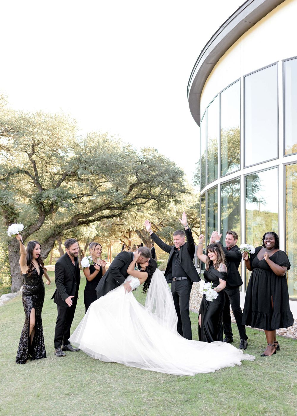 Wedding party in black dresses and suits poses in a curved line against a modern glass building exterior.