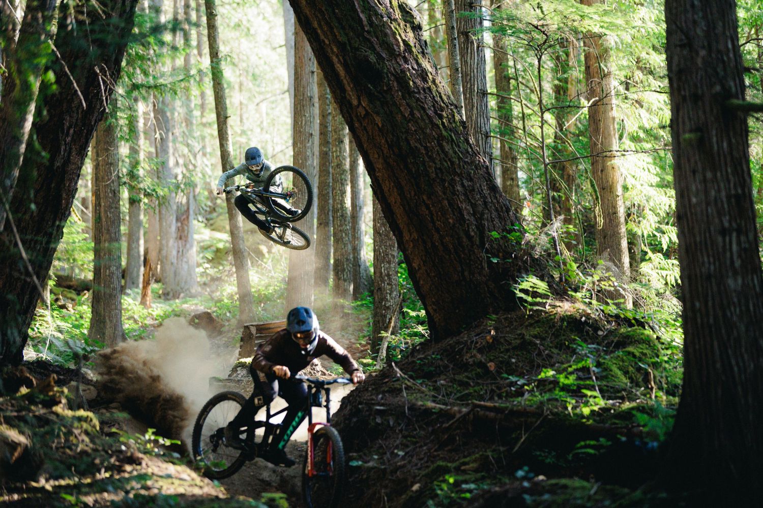 Mountain bikers Caleb Holonko racing through a misty forest trail with Peter Kaiser catching air off a jump.