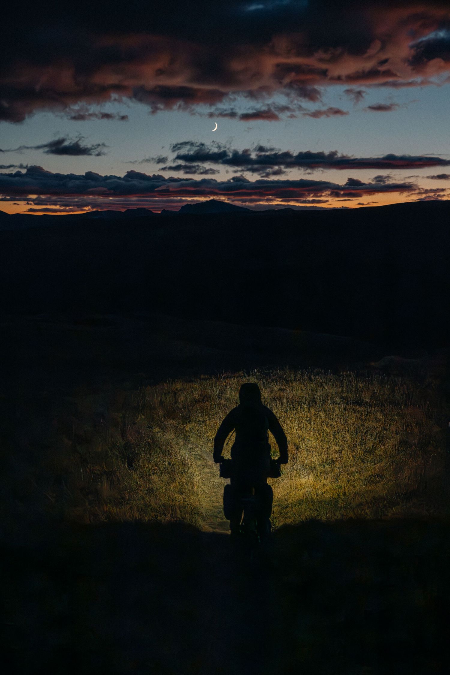 Silhouette of a mountain biker at dusk, headlight illuminating trail, crescent moon visible in dramatic cloudy sky.