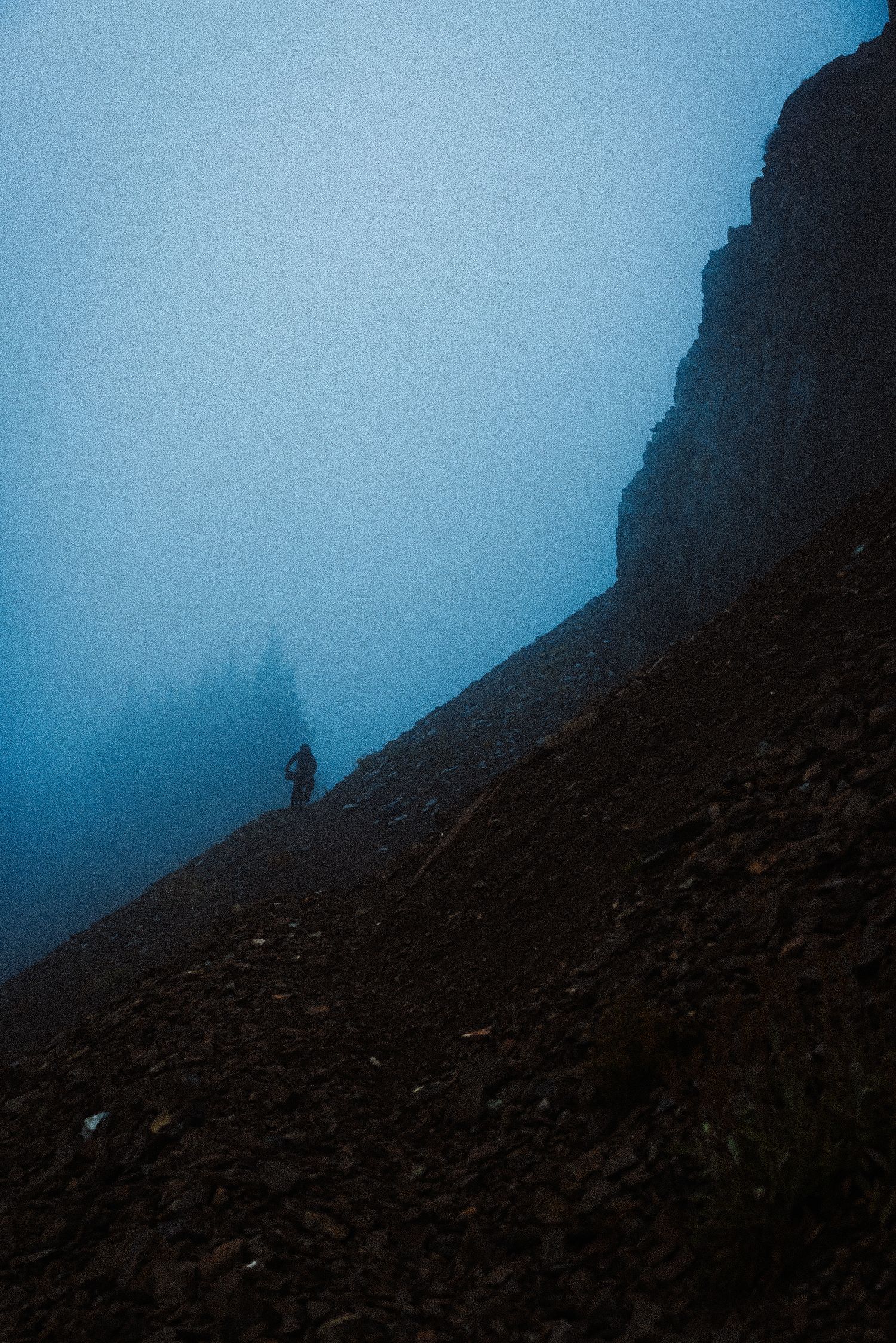 A lone hiker silhouetted against a misty blue mountain trail surrounded by rocky terrain and fog.