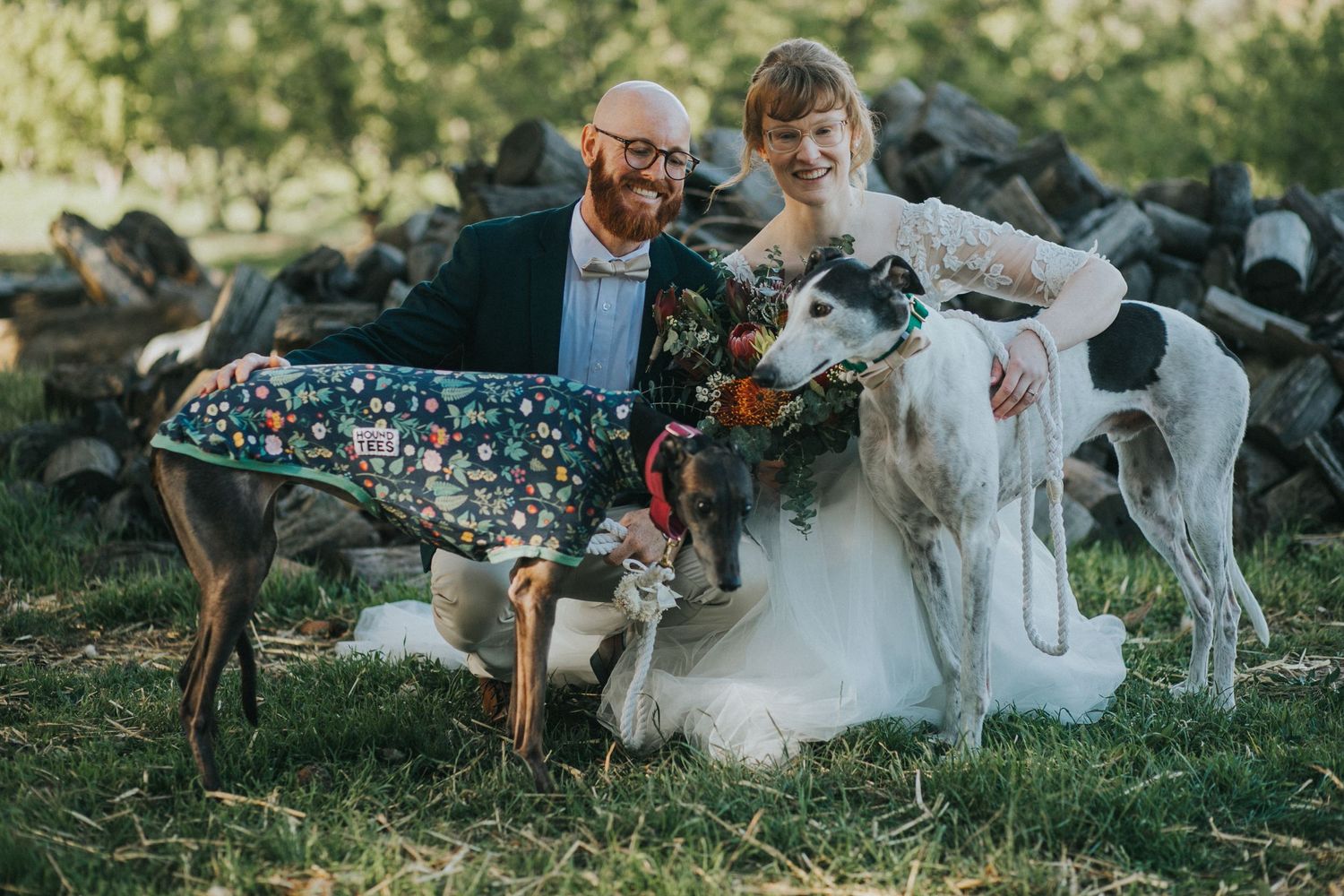 Quirky couple poses with two greyhounds in colorful outfits on grass at outdoor wedding celebration.