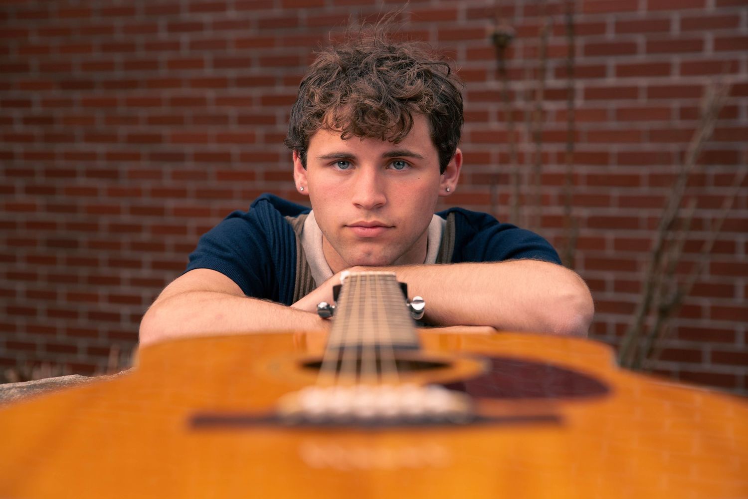 Young man resting chin on acoustic guitar against brick wall background.
