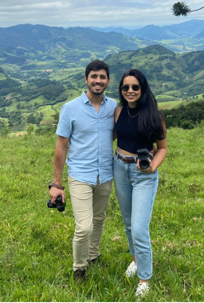 casal sorrindo abraçado em um campo verde com vista panorâmica para montanhas e vales sob um céu azul