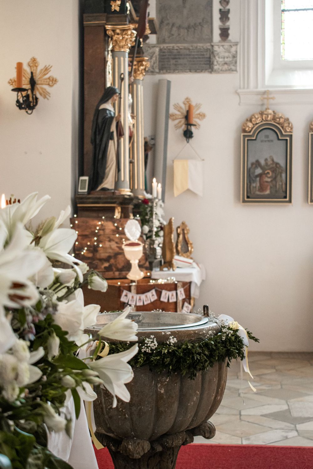 Church interior with baptismal font, white lilies, Virgin Mary statue, and altar decorations for a religious ceremony.