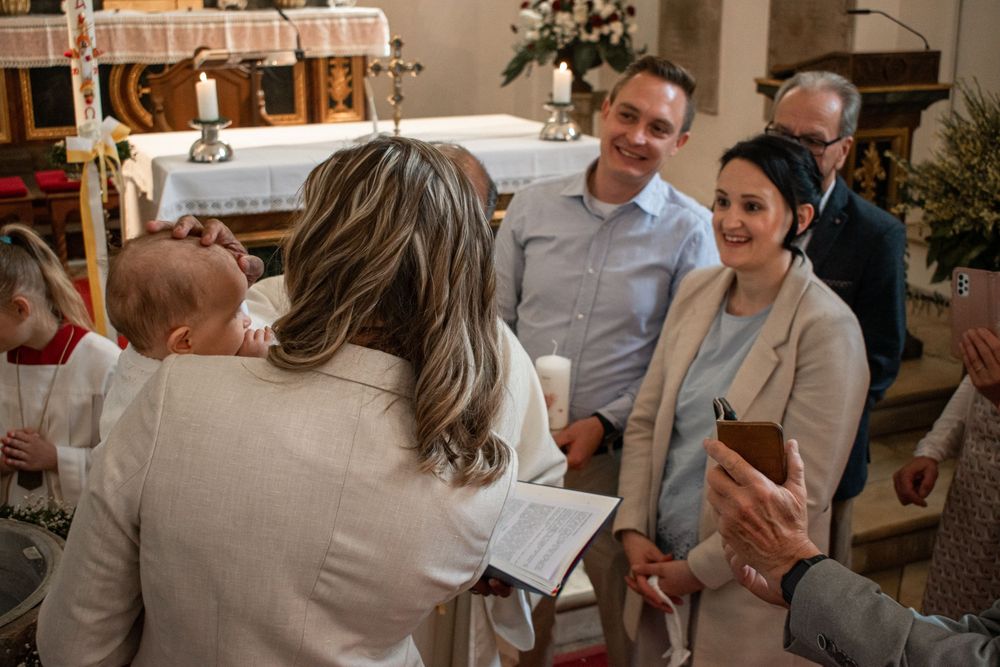 A joyful baptism ceremony inside a church with family members gathered around smiling and holding a baby.