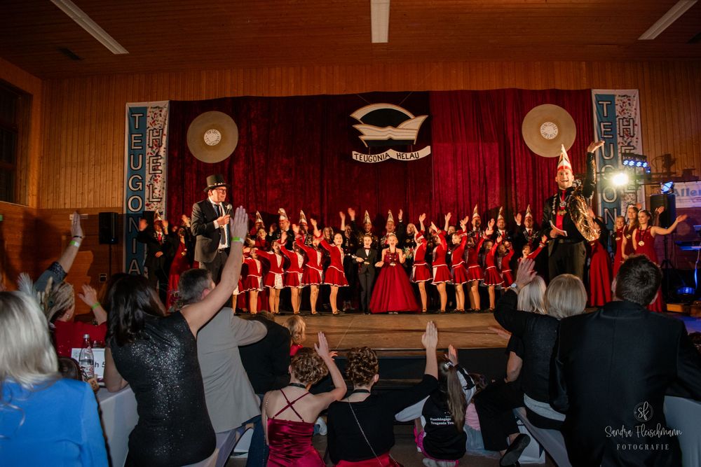 A choir performs on stage in red dresses at a formal concert venue with wood-paneled walls and Christian fish symbol decor.