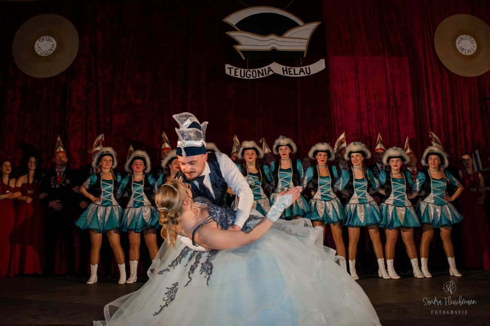 A group of dancers in matching blue costumes perform synchronized movements on a dark stage during a performance.