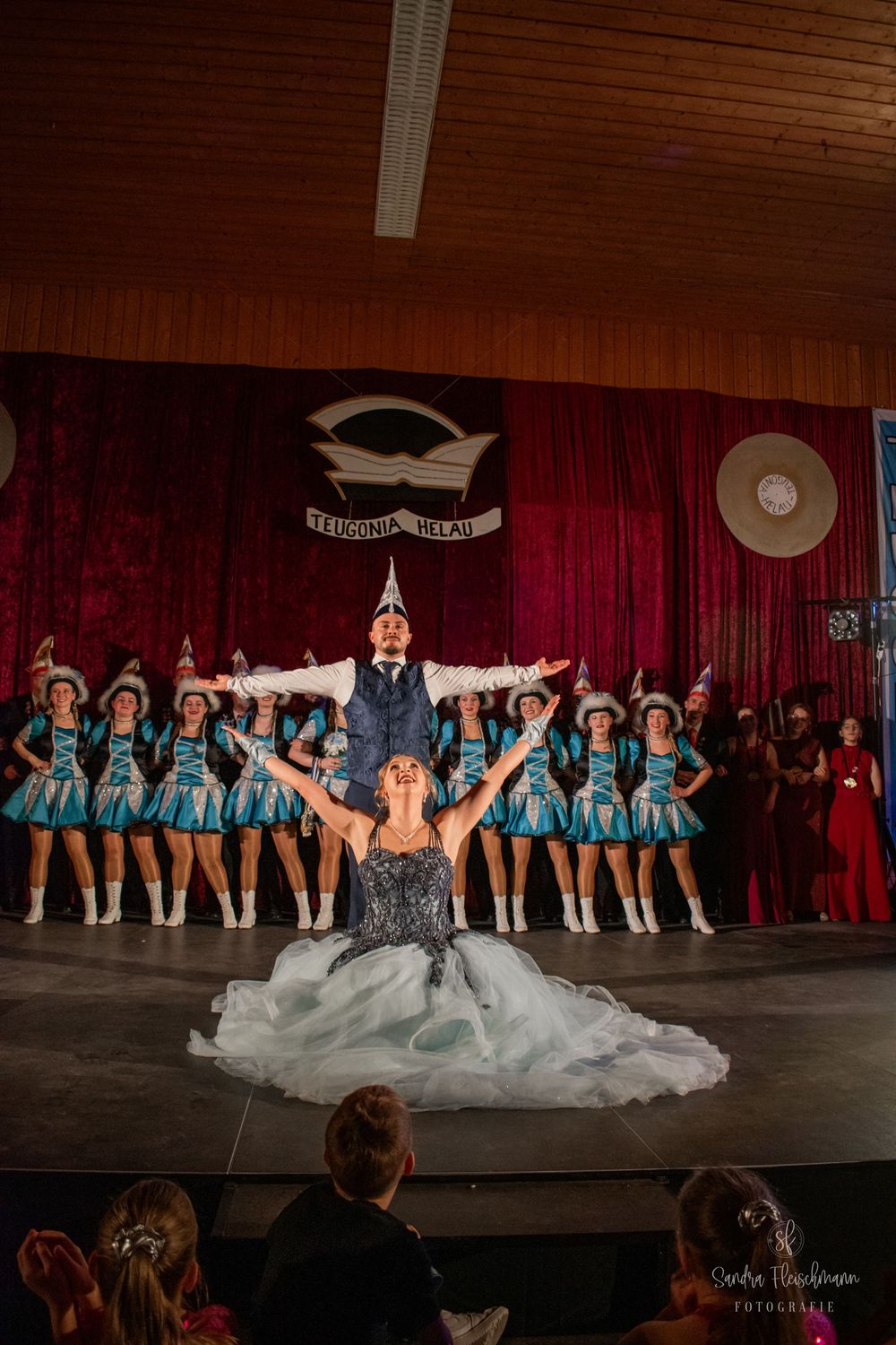 Dancers in matching light blue costumes perform on stage with a central figure in a dramatic flowing gown.