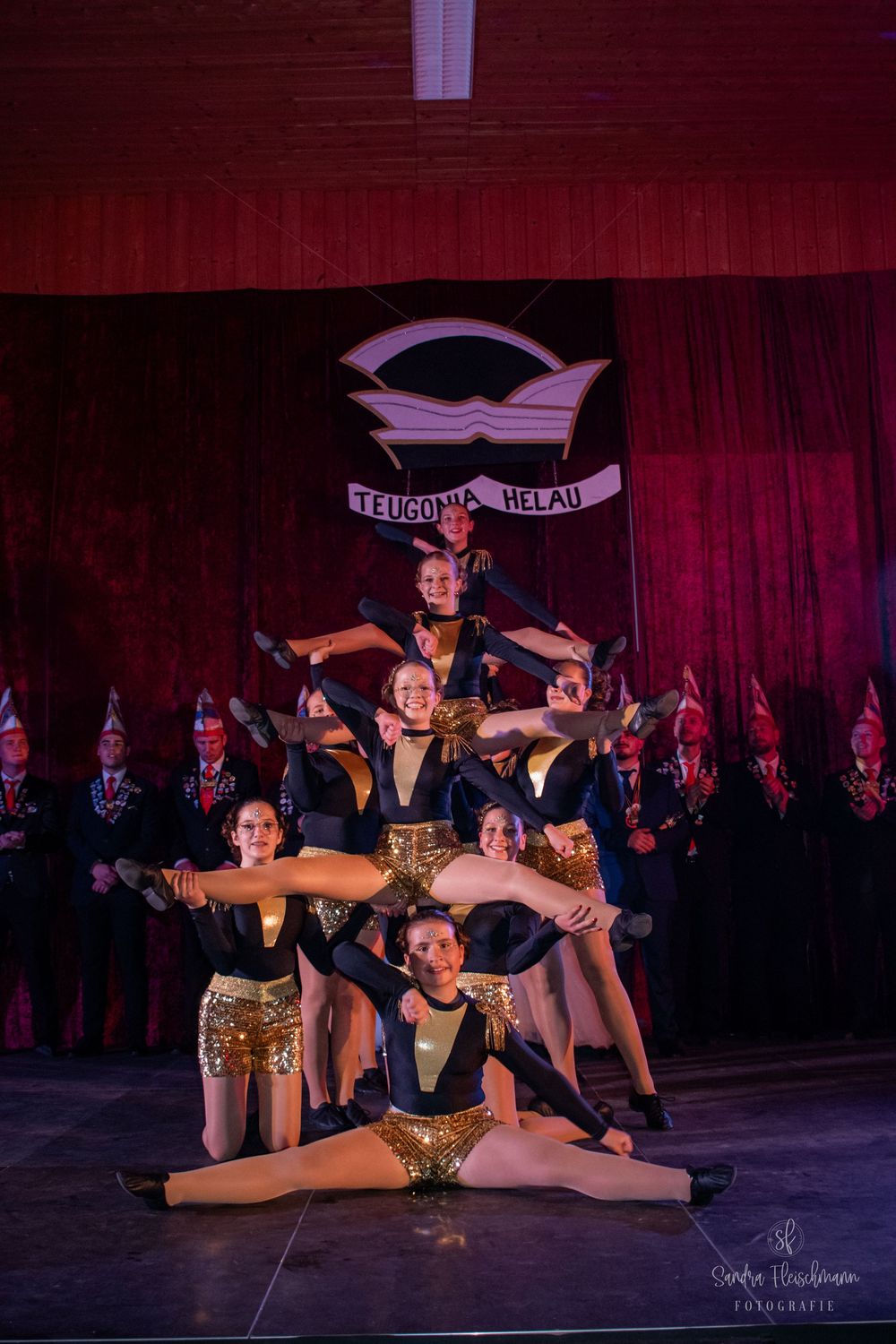 Dancers in gold sequined costumes perform a dramatic split formation on stage with red curtain and choir backdrop.