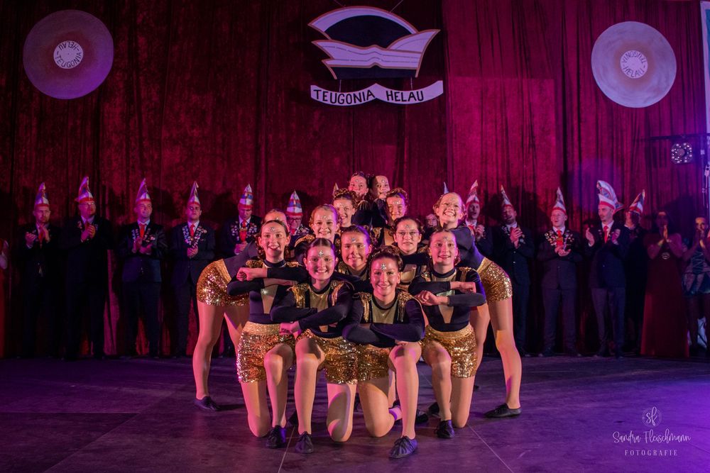 Group of dancers performing in formation on a dimly lit stage with purple lighting effects.