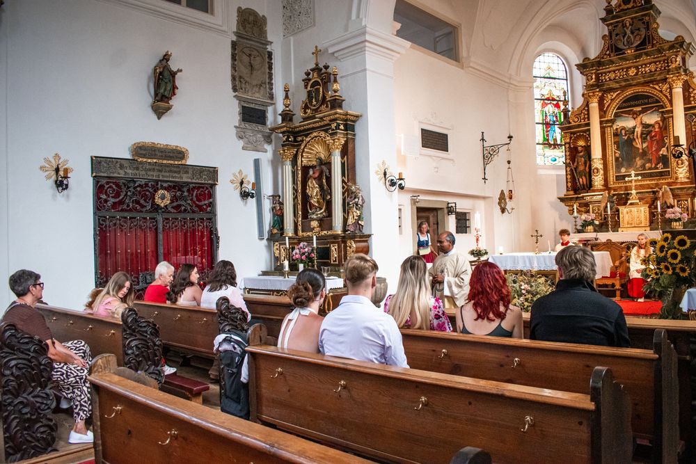 Congregation seated in wooden pews during a Catholic church service with ornate baroque altar and decorations.