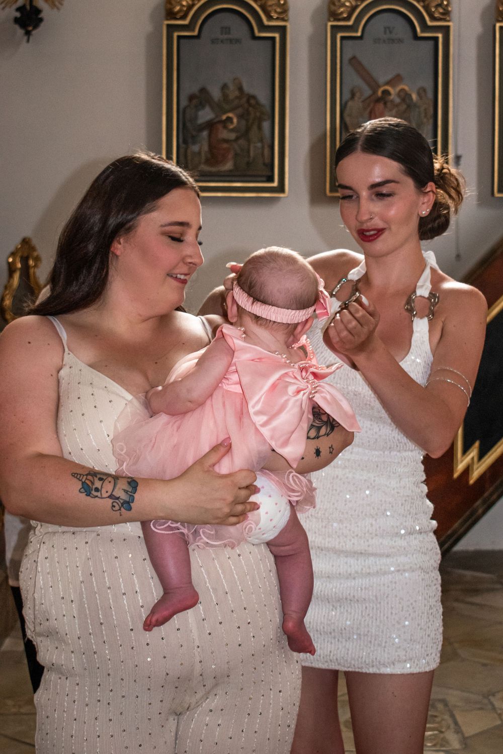 Two women in white dresses admiring a newborn baby in pink outfit inside a church.