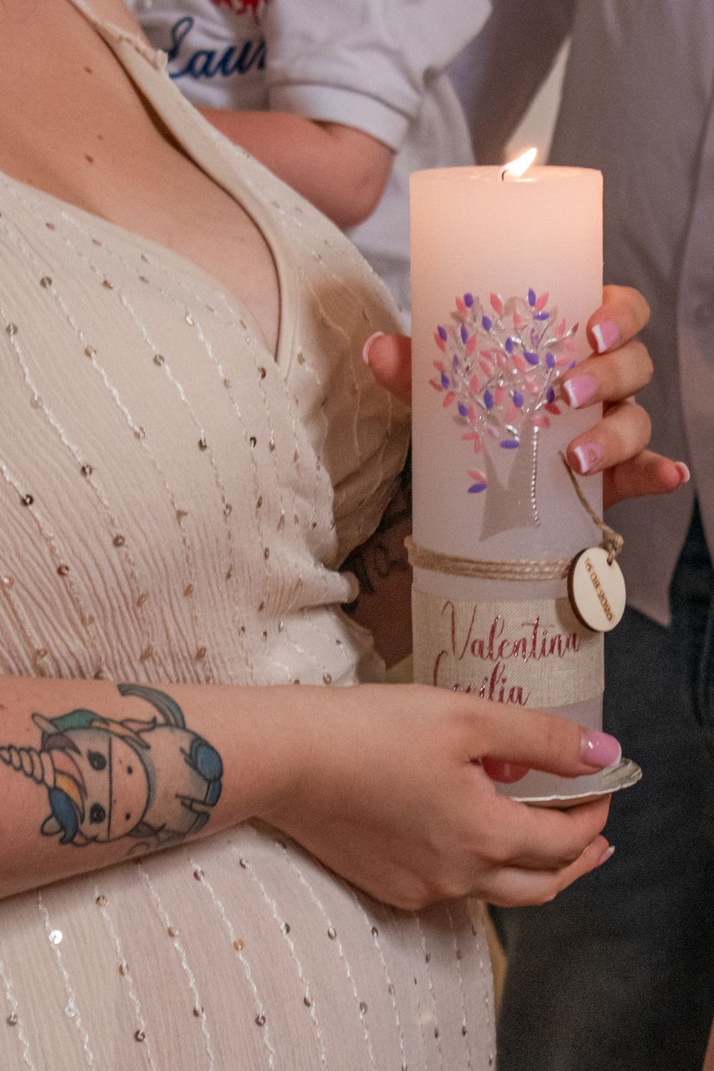 Woman holding a lit personalized baptism candle with a tree design and the name Valentina, wearing a white dress with a unicorn tattoo.