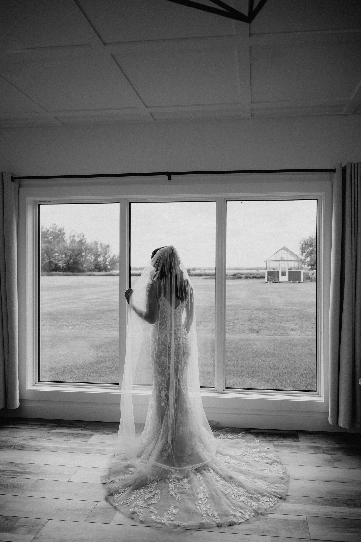 A dreamy silhouette of a bride in white lace dress and veil stands by a window in soft morning light.