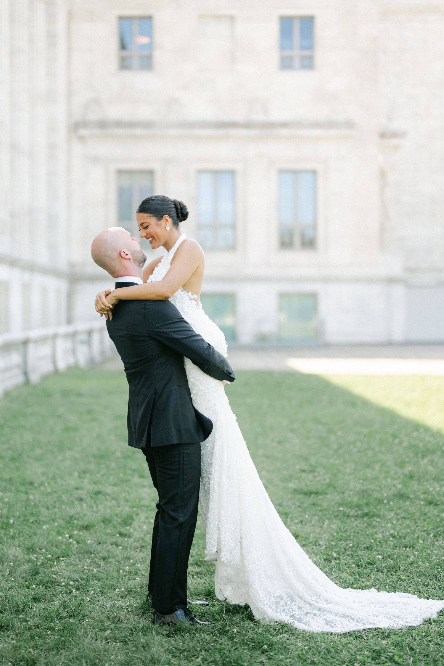 Groom lifts smiling bride in white lace wedding gown outdoors on green lawn beside elegant stone building. - The Field Museum Chicago