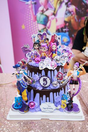 A decorated 5th birthday cake with purple frosting, doll figurines, and colorful cartoon character toppers on a white stand.