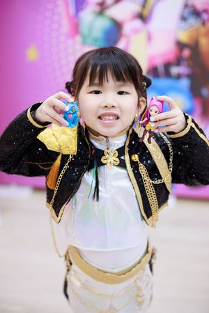 Young Asian girl in sparkly costume holding two small dolls, smiling happily at a colorful event backdrop.