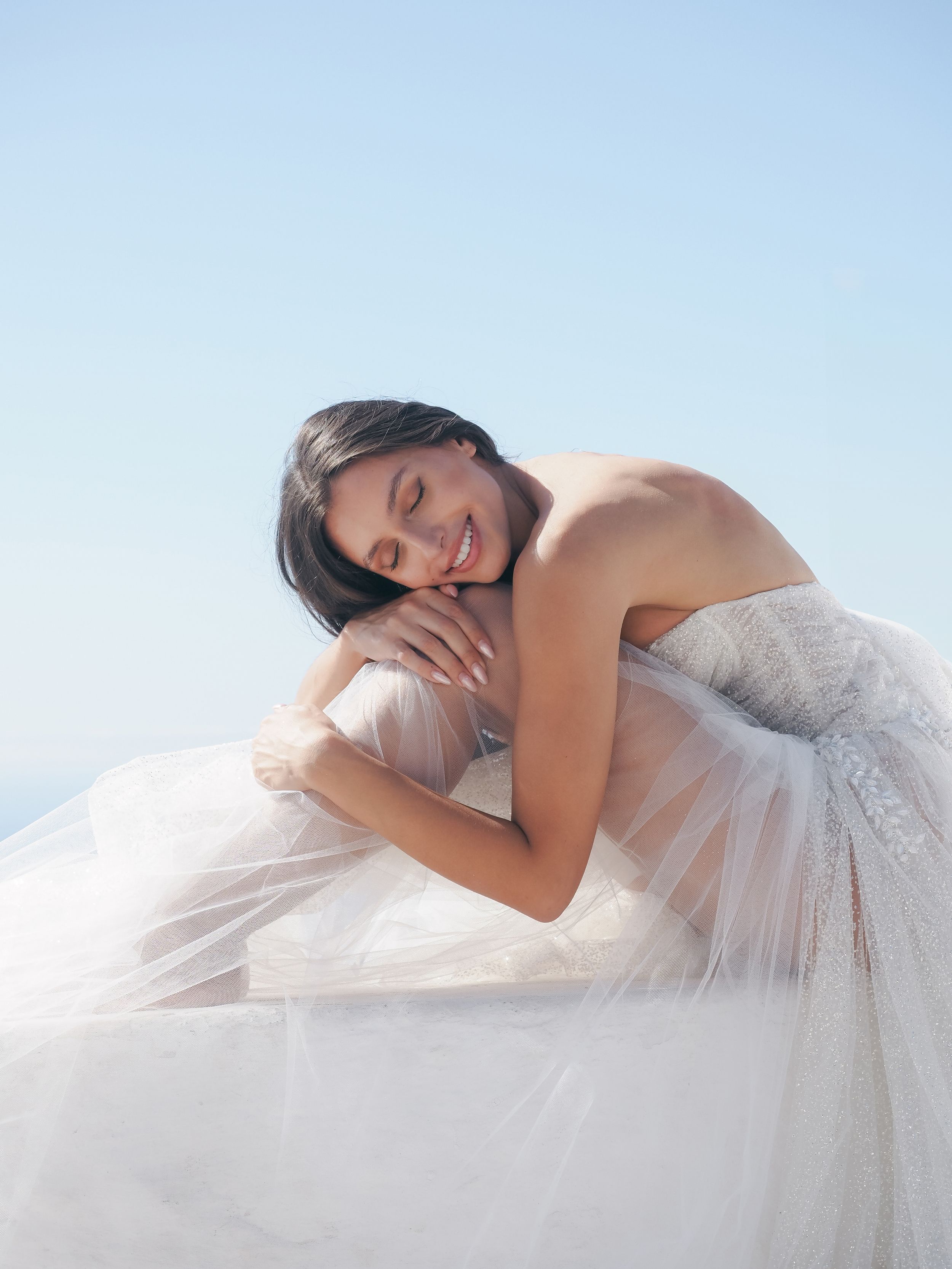 Elegant bridal portrait with flowing white tulle dress against blue sky.