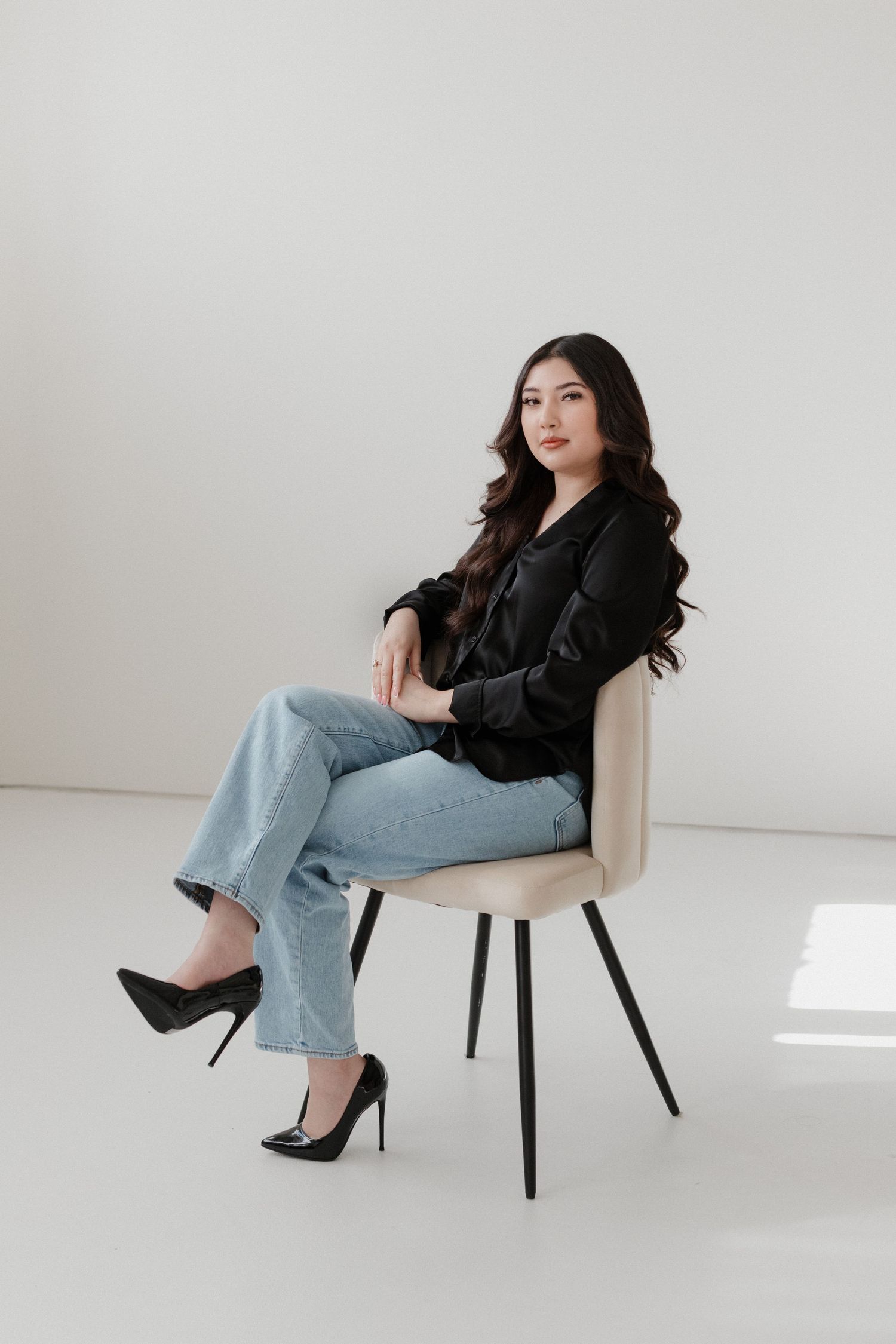 Stylish person poses in chair wearing black blazer, light denim jeans and black stiletto heels against white backdrop.