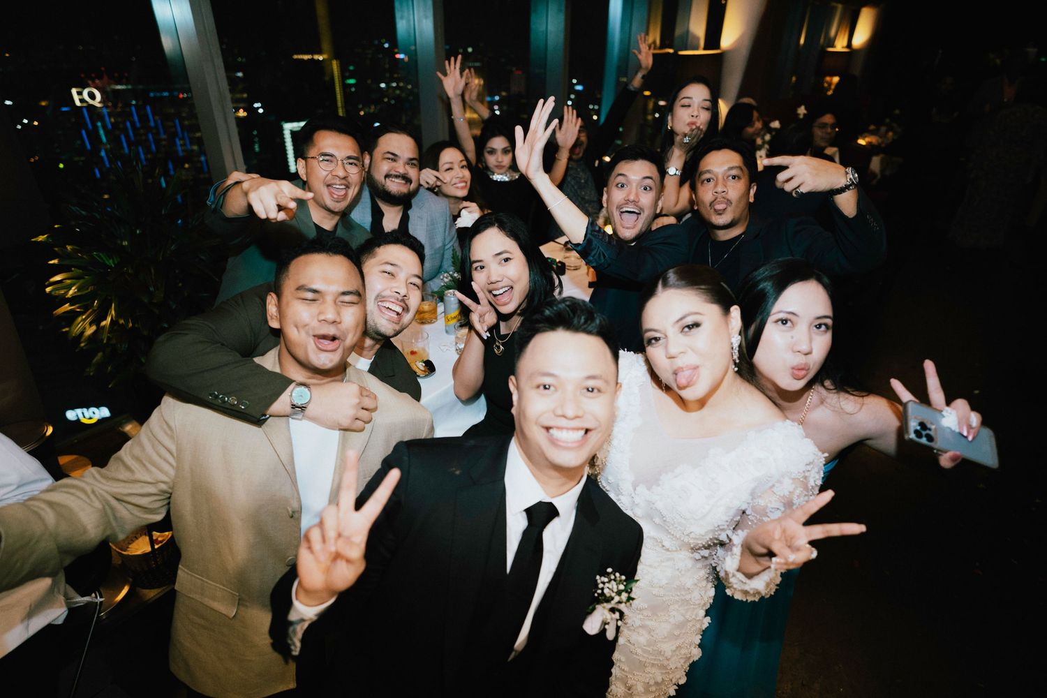 Wedding party group selfie with bride and groom celebrating at a reception venue at night.
