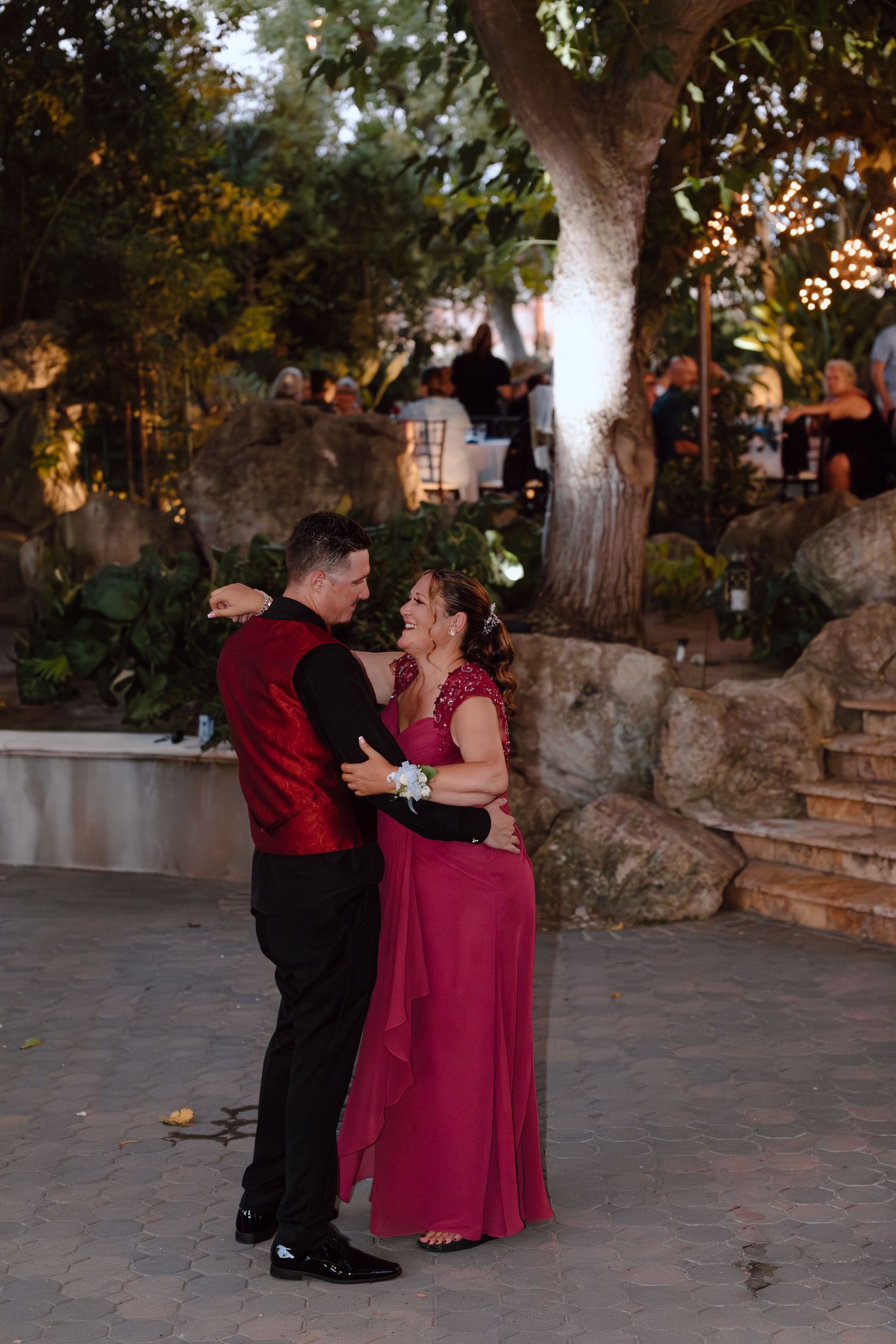 A couple dances together at an outdoor venue with trees and string lights in the background.