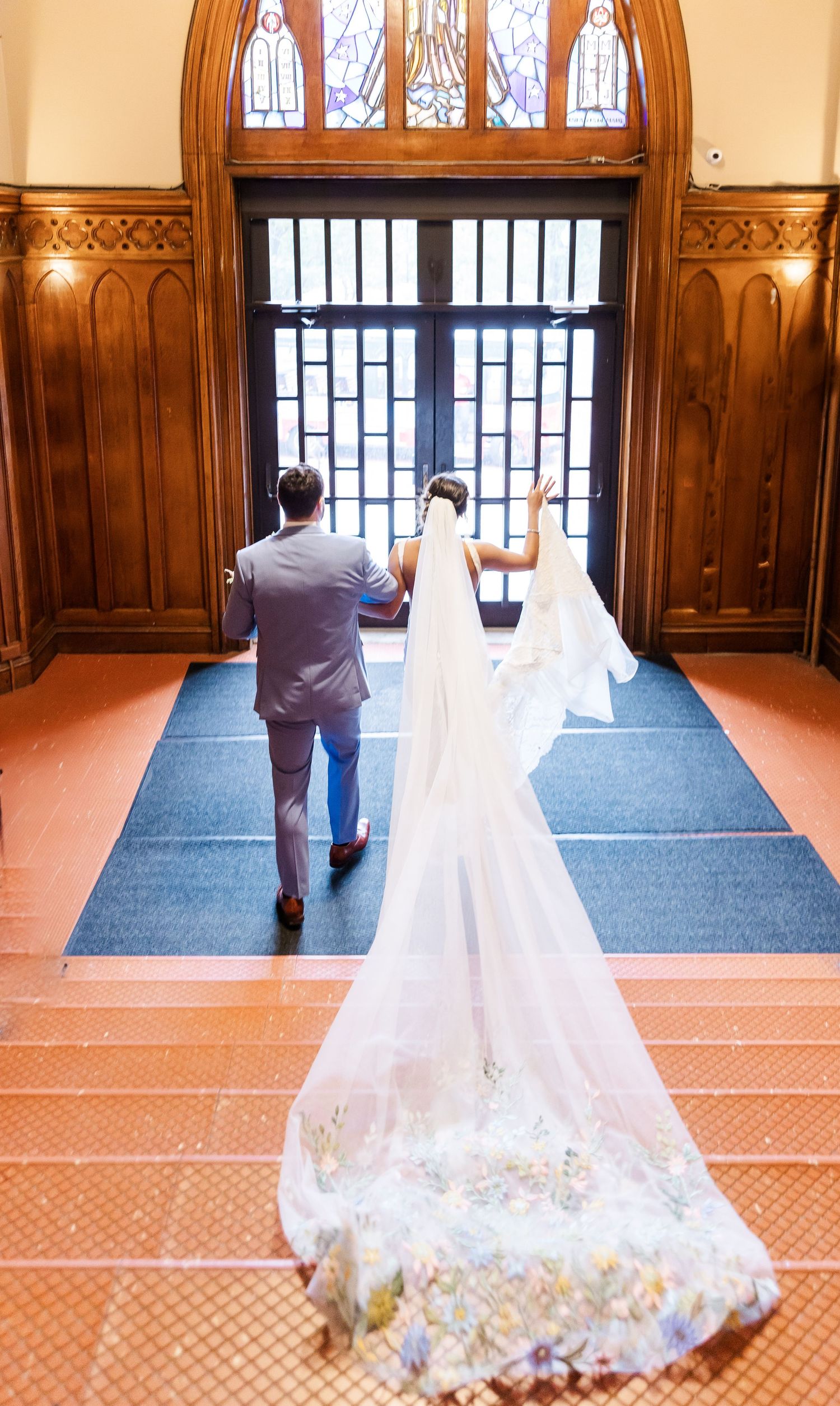 A trailing wedding dress veil follows a couple walking towards grand wooden church doors with arched windows.