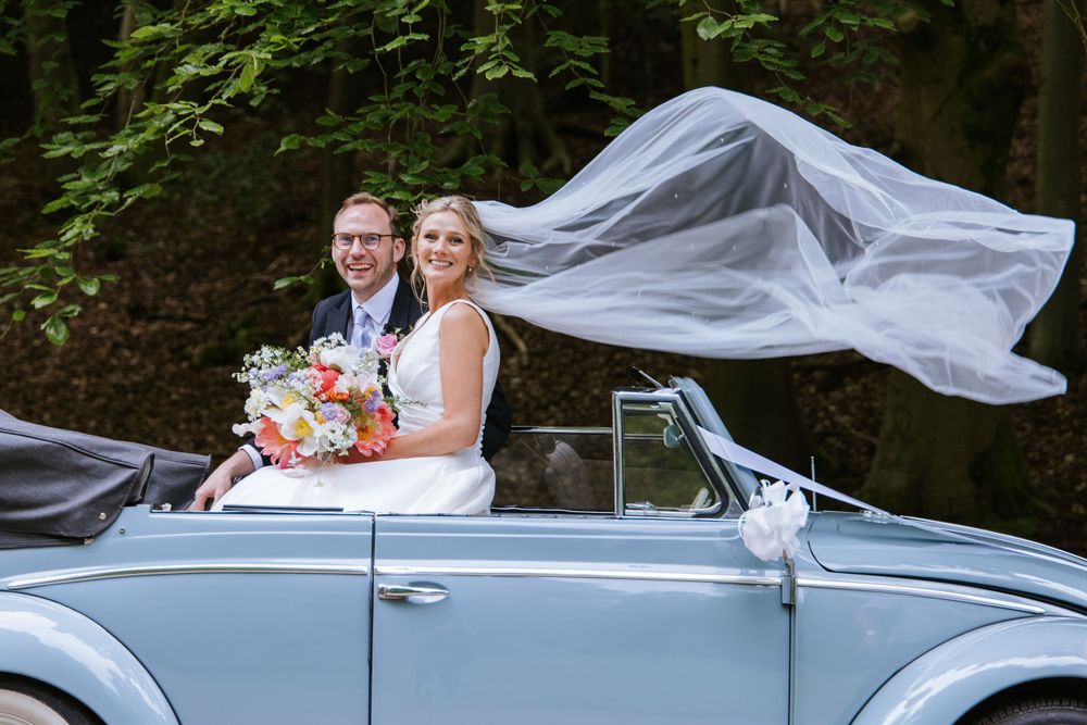 Groom and bride with flowing veil whilst sat in the car on the driveway to Harper Weddings Cain Manor in Hampshire.