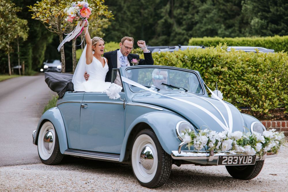 Bride and groom on the driveway to Harper Weddings in the convertible car, both cheering, bride with flowers up high.