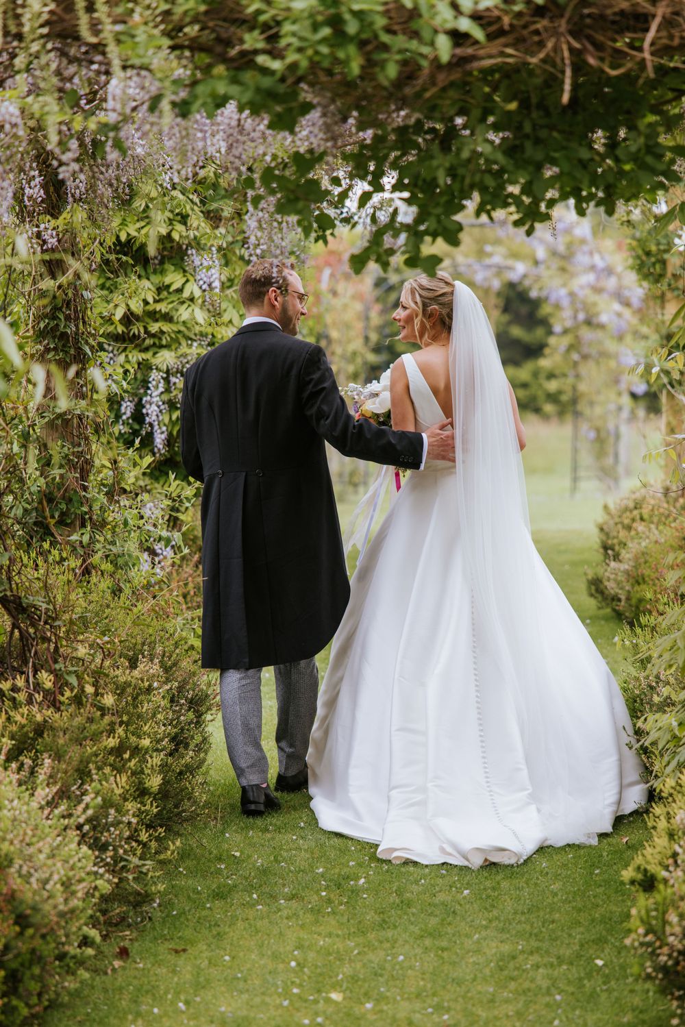 Bride and groom walking through wisteria arch in Harper Weddings garden, in Cain Manor, Hampshire.