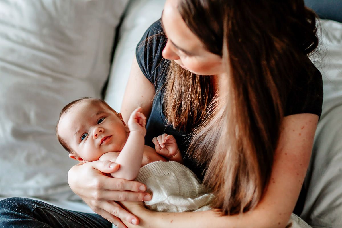 Nyfødtfotografering hjemme hos kunde på Malmheim. Mamma holder nyfødt sønn i fanget og ser ned på han mens de sitter på en seng.