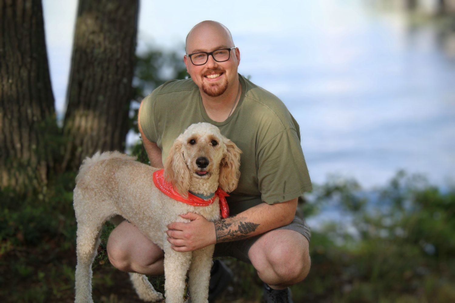 A smiling bald man with glasses hugs a fluffy golden doodle dog outdoors near a lake surrounded by trees.