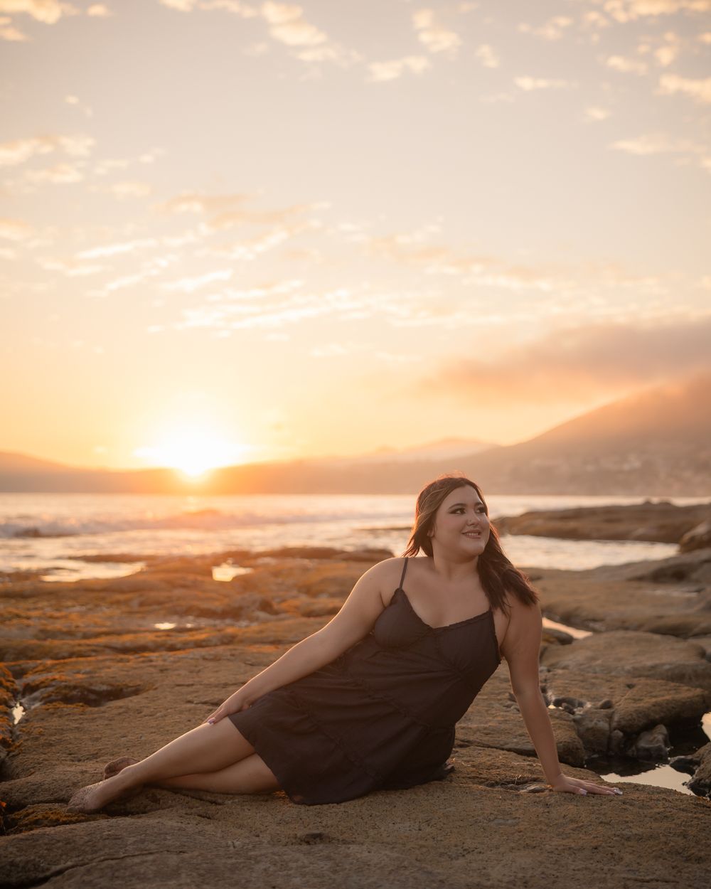 A person poses gracefully on a sandy beach at sunset with rocky coastline and golden sky in the background.