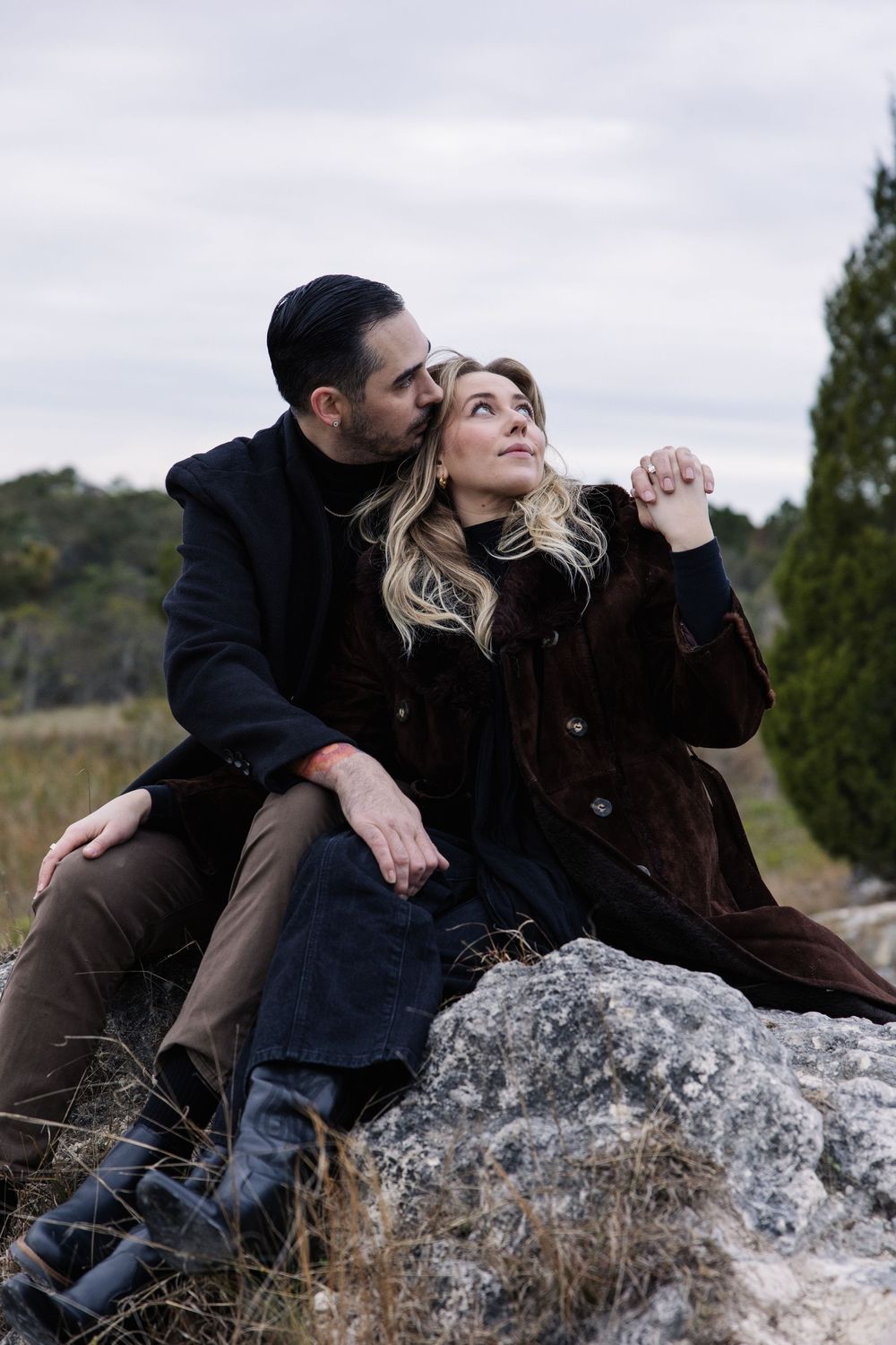 A romantic couple sitting together on a large rock outdoors, sharing an intimate moment against a cloudy sky.