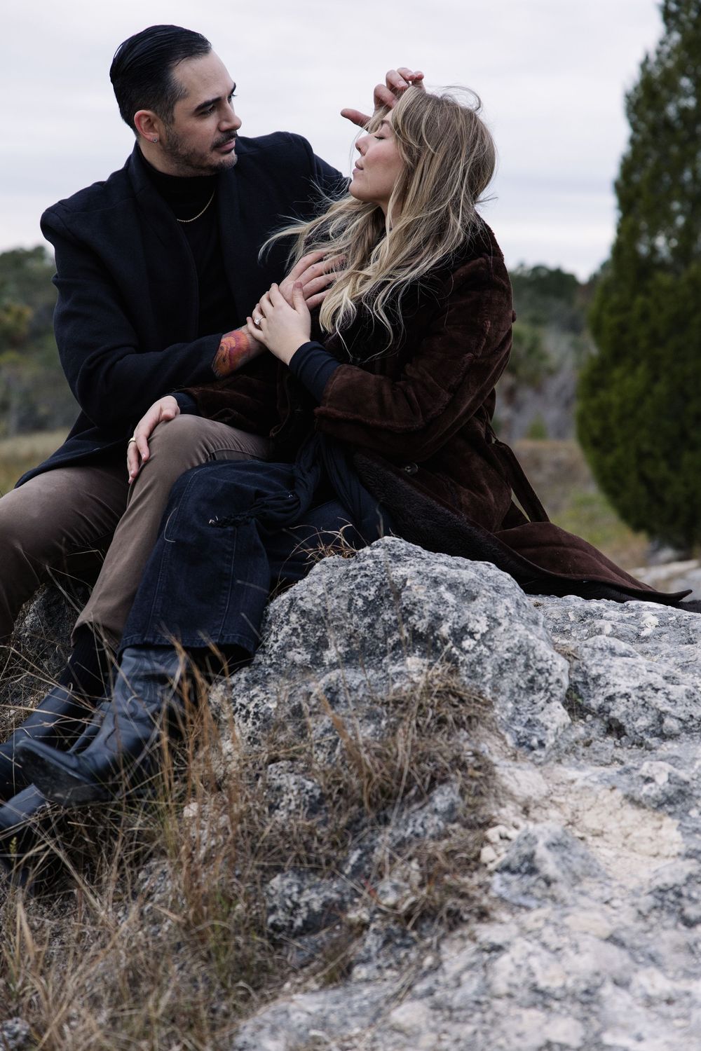 A couple sits intimately together on a rocky outcrop wearing dark winter clothes against an overcast sky.