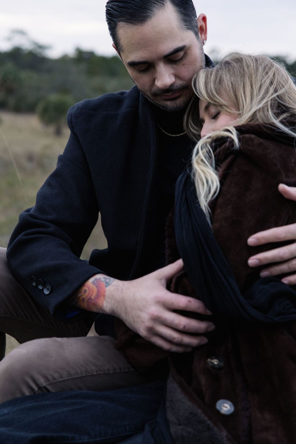 A romantic outdoor portrait shows an intimate embracing couple in dark coats against a blurred natural background.