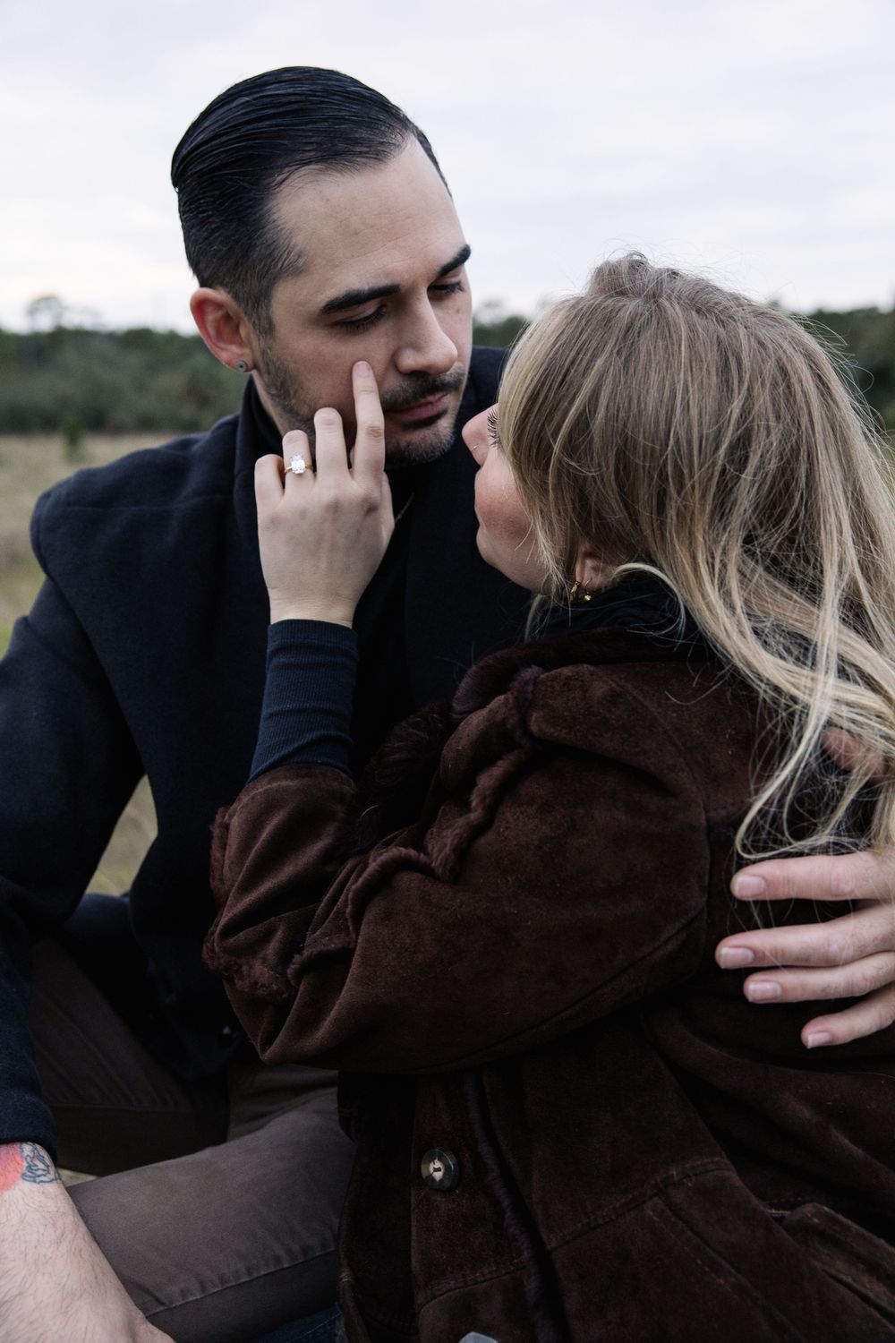 A romantic couple share an intimate moment outdoors, with the woman in a brown coat touching the man's face tenderly.
