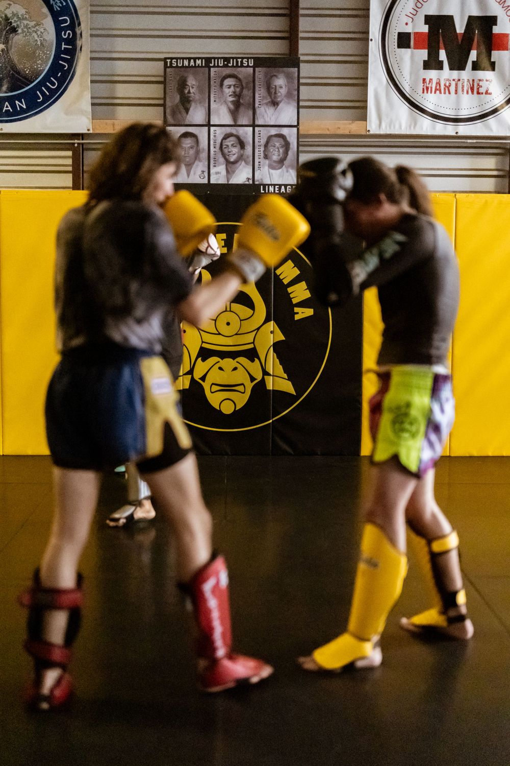 Two martial artists spar in front of yellow wall with samurai logo at training facility.