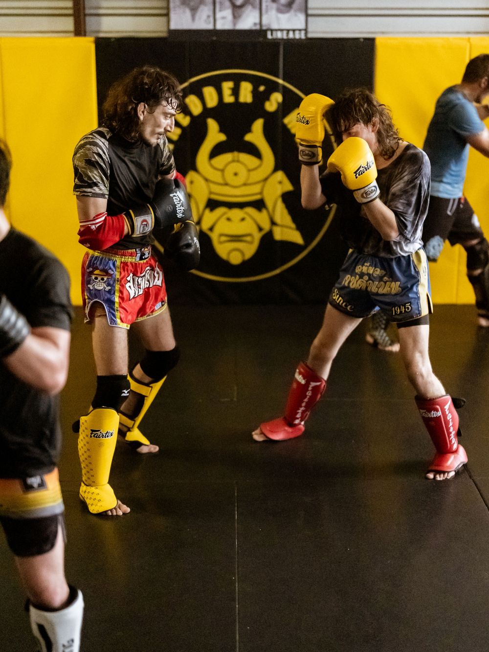 Fighters in colorful gear sparring in front of black and yellow gym wall with samurai logo.
