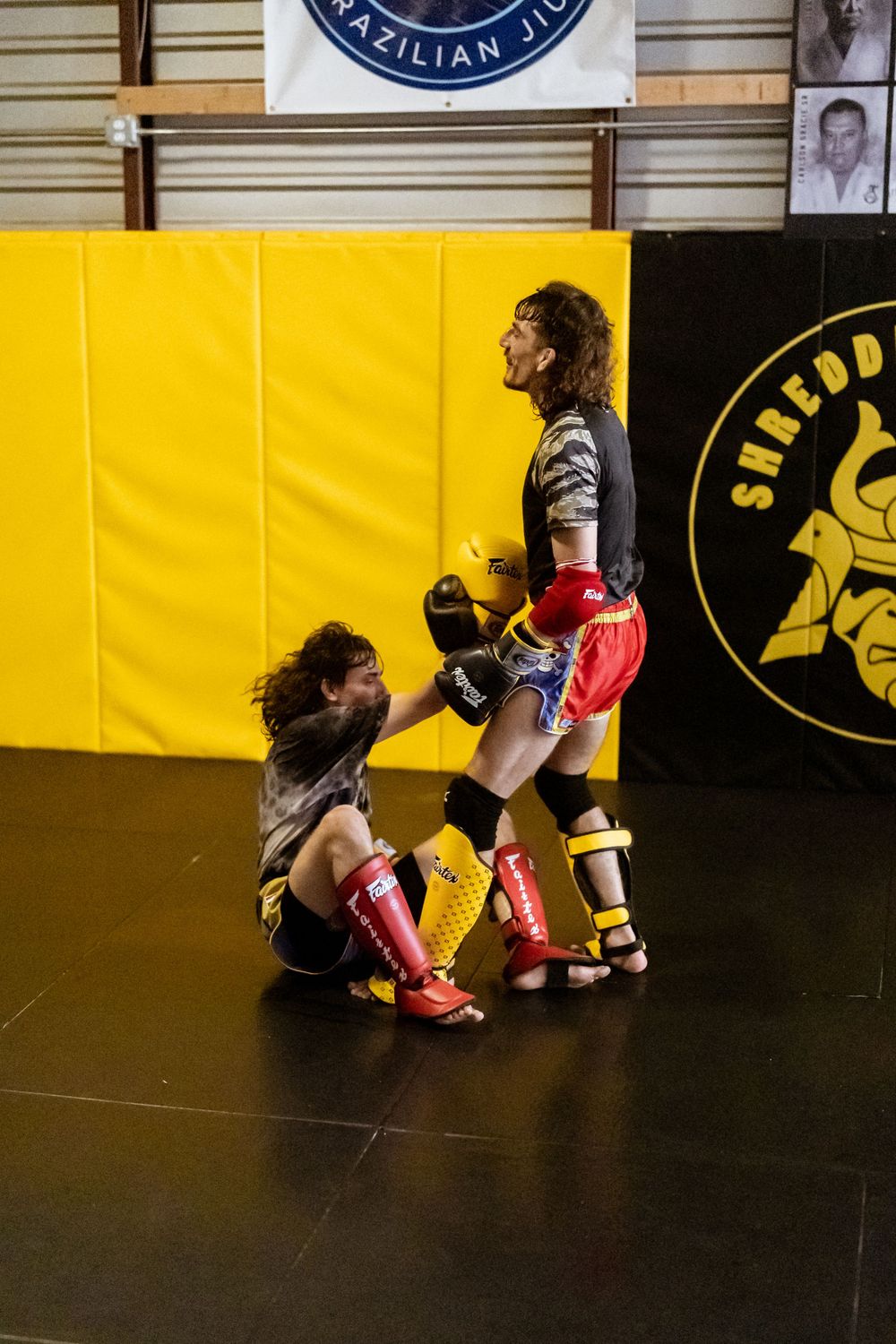 Two fighters practicing kickboxing moves in a martial arts gym with yellow padded walls and training logos.