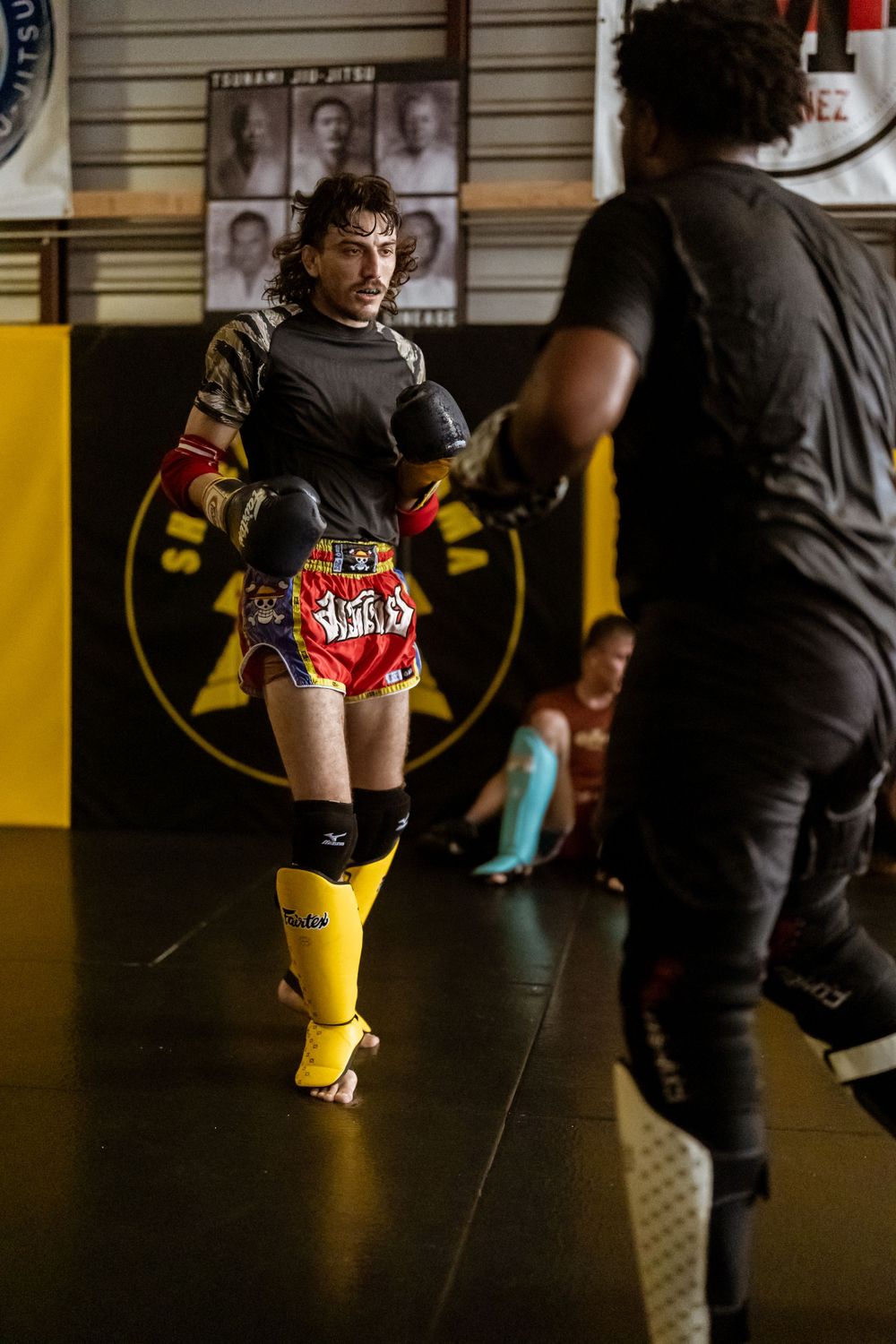 Martial artist in yellow boots and shorts training with partner in a combat sports gym.