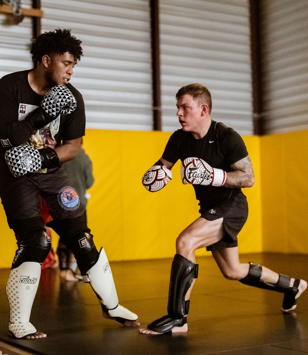 Two fighters practicing combat techniques wearing protective gear in a training facility.