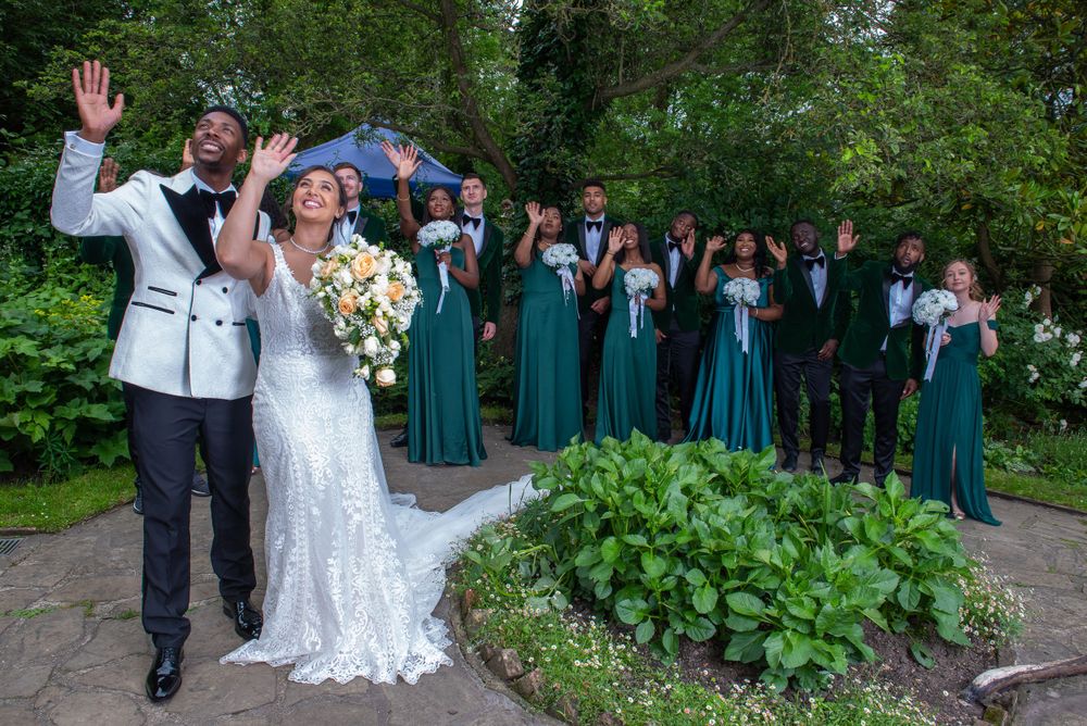 Wedding party in emerald green dresses poses on garden path with celebratory gestures.