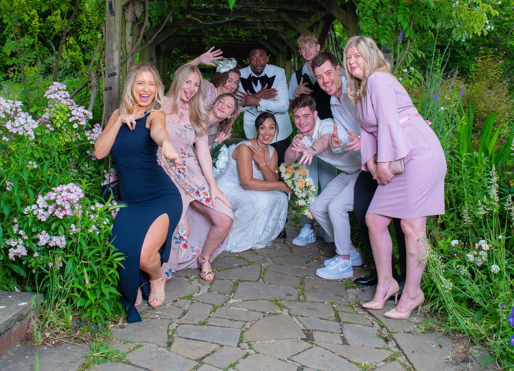 A bridal party poses together outdoors on a stone path surrounded by blooming pink flowers and greenery in a garden setting.