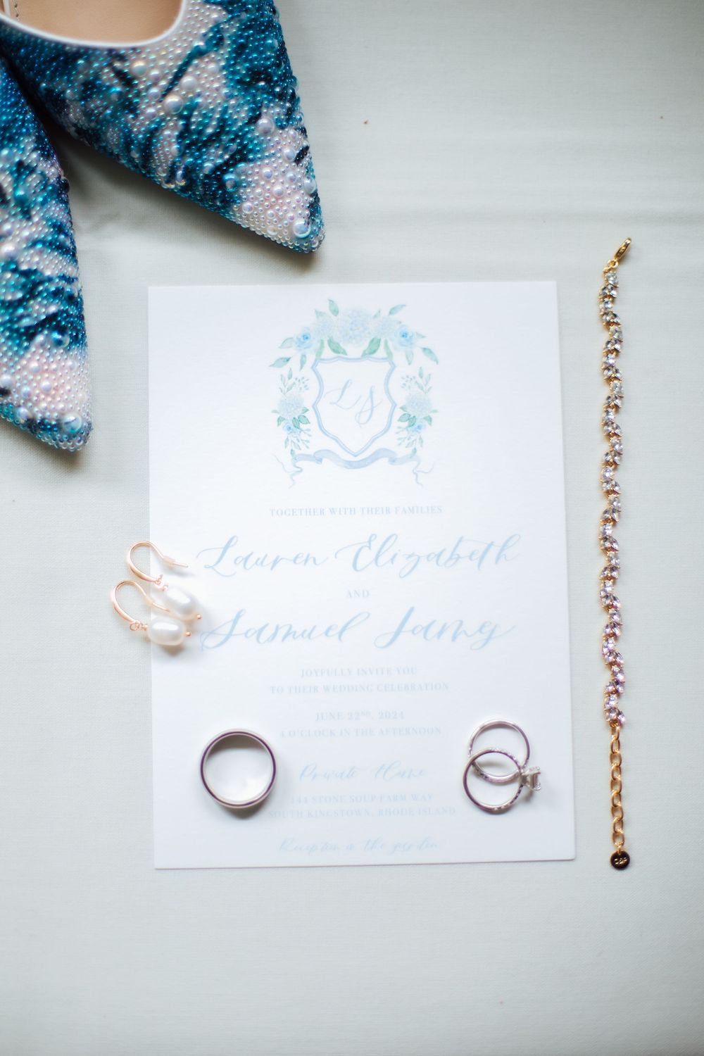 Wedding flat lay featuring blue beaded heels, gold bracelet, rings, pearl earrings, and elegant watercolor monogram invitation.