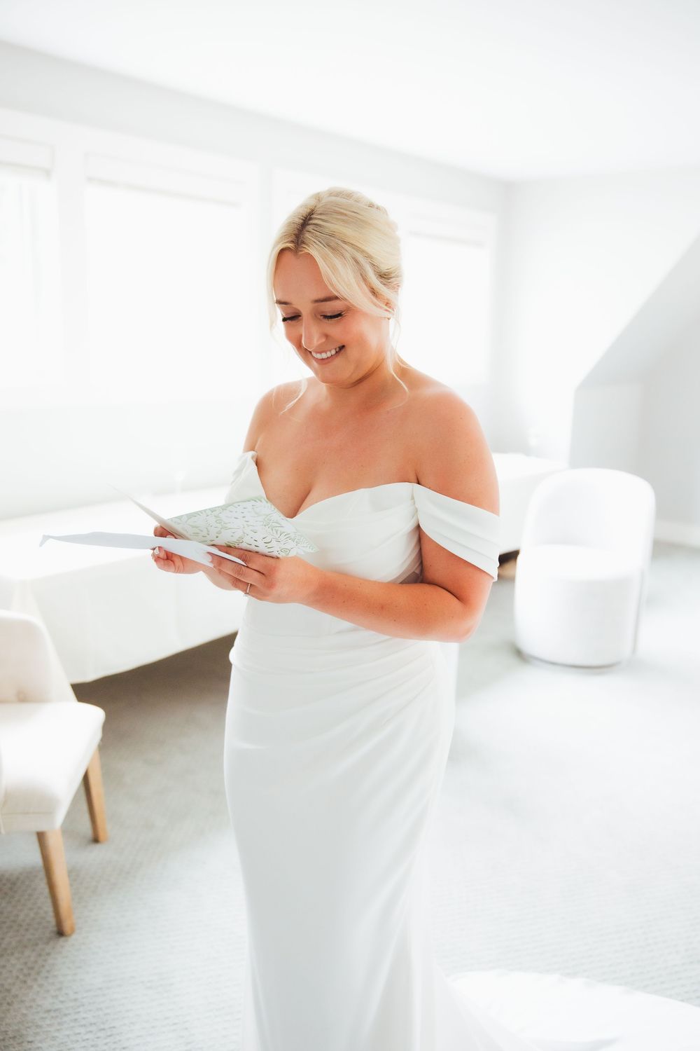 Smiling bride in off-shoulder white gown reading a card in a bright, modern room on her wedding day.
