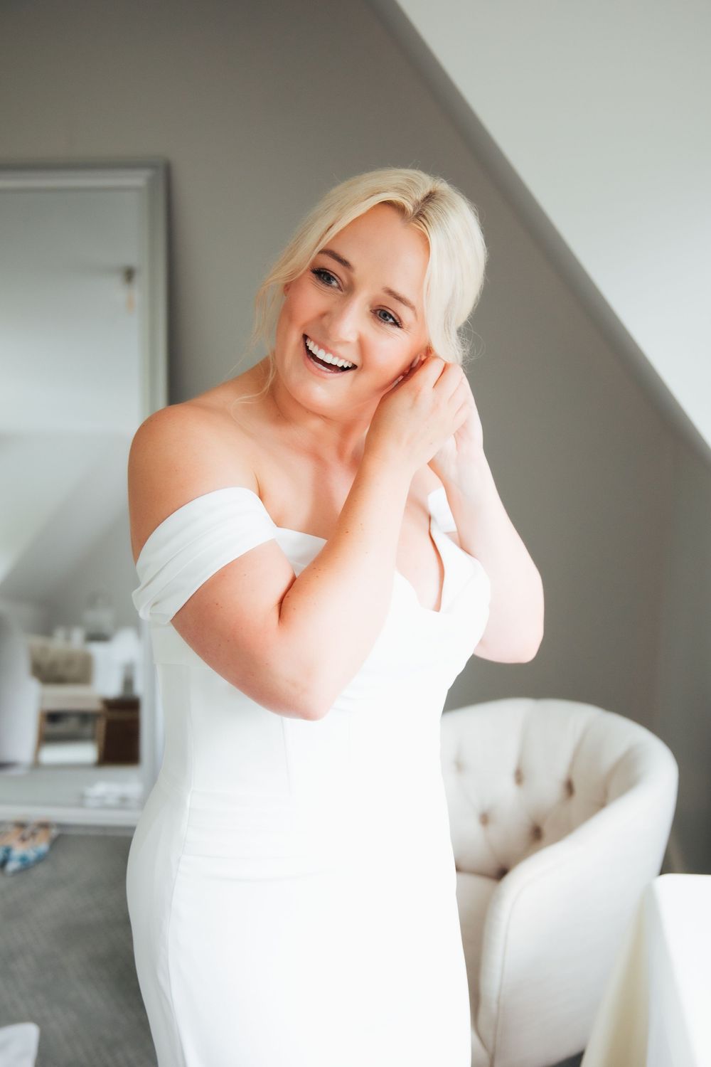 Smiling blonde bride in an off-shoulder white wedding dress posing in a bright room.