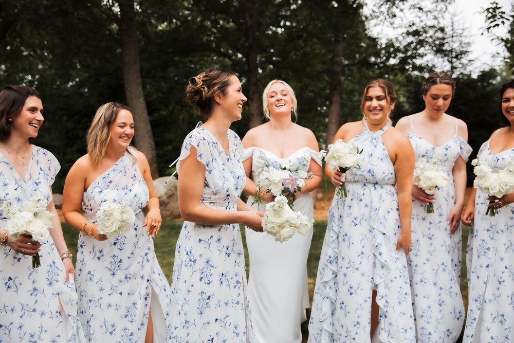 Bride and bridesmaids laughing together in matching blue floral dresses, holding white bouquets outdoors.